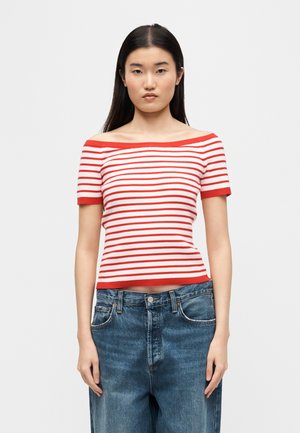 Femeie cu păr lung și închis la culoare, purtând un top roșu cu dungi albe, fără umeri și cu mâneci scurte, și blugi lejere albaștri, stând în picioare pe un fundal simplu.