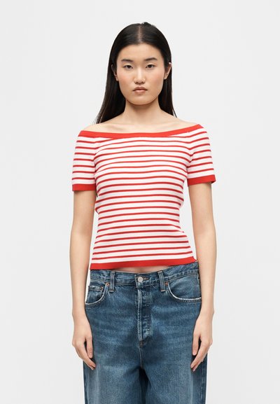 Γυναίκα με μακριά σκούρα μαλλιά που φοράει κόκκινη και λευκή ριγέ μπλούζα με κοντά μανίκια και έξω τους ώμους, και άνετο μπλε τζιν, στέκεται μπροστά σε απλό φόντο.