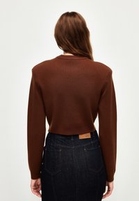 Femme aux longs cheveux bruns portant un haut marron à manches longues et un jean taille haute bleu foncé, vue de dos sur un fond uni.
