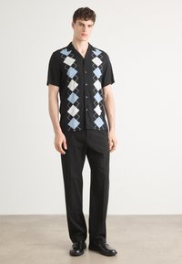 Chemise noire à manches courtes avec un motif losange bleu et blanc, boutons et col, associée à un pantalon noir et des chaussures noires.