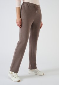 Pantalones de chándal marrones, elásticos, con cintura ajustable y bolsillos laterales, combinados con zapatillas blancas con detalles dorados.
