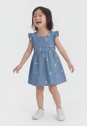 Mladé dievča sa usmieva, má na sebe modré bezrukávové šaty s farebnými vzormi zmrzliny a srdiečok a biele tenisky, stojí s otvorenými rukami.