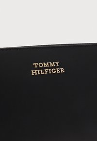 Melnas ādas maks ar teksturētu virsmu un zelta "TOMMY HILFIGER" logotipu, kas izcili redzams uz priekšpuses.