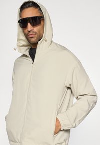 Világos bézs cipzáras kapucnis dzseki, rugalmas mandzsettákkal és két oldalzsebbel. Fekete ing fölött viselve, sötét napszemüveggel párosítva.