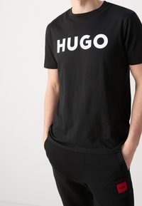 Camiseta de algodón negra con un gran logo blanco "HUGO" en el frontal, diseño de cuello redondo y mangas cortas, combinada con pantalones negros.