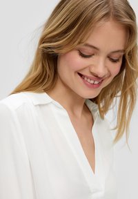 Balta blusa su V formos iškirpte, minkšta medžiaga ir subtili tekstūra. Turi ilgus rankovės ir klasikinį apykaklę. Sklandi, lengva drapiruotė.