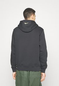 Sudadera negra con capucha y un logo de Nike amarillo en la parte trasera, hecha de una tela suave con un corte relajado. Combinada con pantalones cargo verdes.