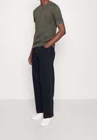 Olivengrön mönstrad kortärmad stickad skjorta i kombination med mörkblå avslappnade jeans och vita sneakers, vilket framhäver en avslappnad stil och komfort.