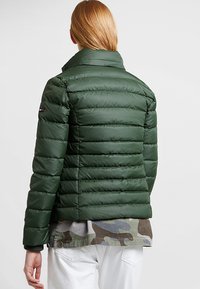 Grön vadderad jacka med krage, horisontell sömnad och elastiska muddar. Bärs över en skjorta med kamouflagemönster, med en slät textur.