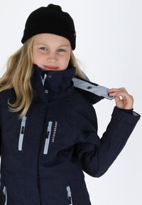 Granatowa kurtka na zewnątrz z wysokim kołnierzem, zapinana na zamek, z kieszeniami na zamek i jasnoniebieskimi akcentami. Czarna dziana czapka uzupełnia całość.