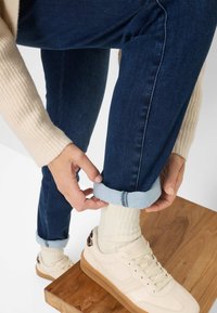 Jeans in denim blu scuro con una risvolto in denim blu chiaro. È visibile un maglione lavorato a maglia color beige chiaro, calzini color crema e una sneaker beige con suola marrone.