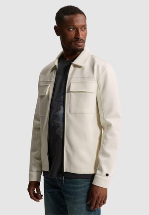Homme portant une veste crème à fermeture éclair sur une chemise noire et un jean bleu, regardant vers sa gauche sur un fond uni.