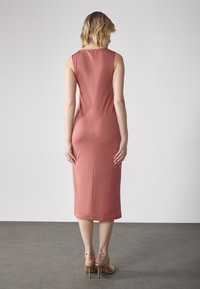 Robe midi ajustée en rose poussiéreux, fabriquée en tissu lisse, avec un col rond et sans manches. Associée à des sandales à talons dorés.