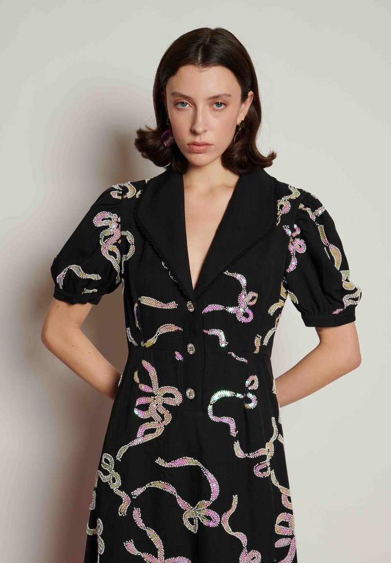 Μαύρο φόρεμα με βαθύ V-λαιμόκοψη, κοντά φουσκωτά μανίκια και μεταλλικές διακοσμήσεις σε κυκλωτά σχέδια. Διαθέτει διακοσμητικά κουμπιά και υφάσμα με υφή.