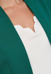 Vihreäkiiltoinen bleiseri, jossa on ribbineulos, yhdistettynä valkoiseen toppiin, jossa on aaltoileva kaula-aukko. Painotus kankaassa ja selkeässä värikontrastissa.