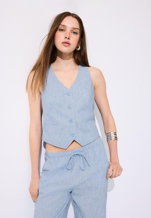 Giovane donna che indossa un gilet azzurro chiaro con bottoni e pantaloni coordinati con coulisse, posando davanti a uno sfondo bianco con un bracciale a polsino.