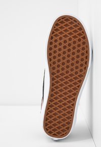 Semelle de chaussure avec motif gaufré en caoutchouc marron sur une semelle intermédiaire blanche, positionnée verticalement contre des murs d’angle blancs.