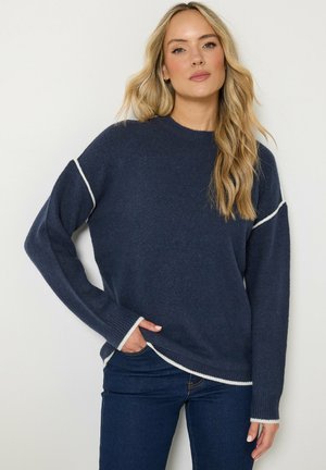 Blonde Frau trägt einen dunkelblauen Pullover mit weißem Rand und dunkelblaue Jeans, steht mit einer Hand in der Tasche vor einem einfarbigen Hintergrund.