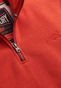 Sudadera roja con cremallera parcial, hecha de material suave, que presenta una textura en el tejido y un logo bordado en el pecho.