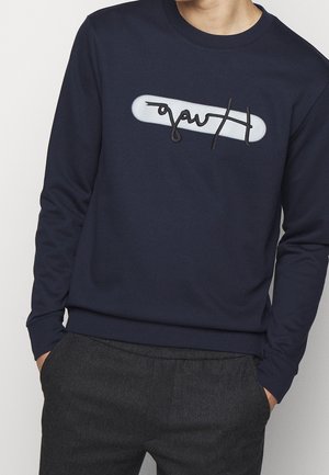 Homme portant un sweat-shirt bleu marine avec un graphisme ovale blanc et un texte noir de style manuscrit sur la poitrine, associé à un pantalon gris foncé.