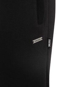 Detail černé tkaniny se dvěma štítky značky SMILODOX přišitémi poblíž švu boční kapsy.