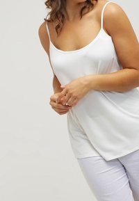 Valkoinen toppi ohuilla ohuilla olkaimilla, pehmeää kangasta, hieman istuva muotoilu ja pyöreä pääntaite, yhdistettynä vaaleisiin housuihin.