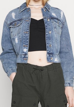 Personne portant une veste en jean bleu délavé par-dessus un crop top noir et un pantalon cargo vert foncé, les mains dans les poches.