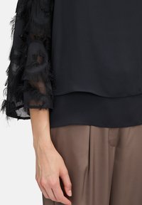 Blusa nera con maniche trasparenti e dettagli in piuma, design a strati e una texture di tessuto liscia. Indossata con pantaloni beige.