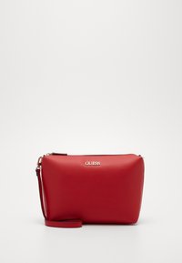 Červená syntetická kožená taška přes rameno s hladkým povrchem, zipovým uzávěrem a odnímatelným popruhem. Na přední straně se nachází lesklé logo značky.