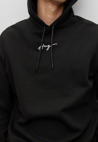 Sweat à capuche en coton noir avec un cordon de serrage, présentant un motif de texte brodé en blanc sur la poitrine. Texture douce et coupe standard.