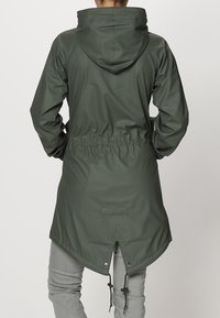 Personne portant un imperméable à capuche vert olive avec taille cintrée et ourlet en queue de poisson, associé à un pantalon gris clair, de dos.