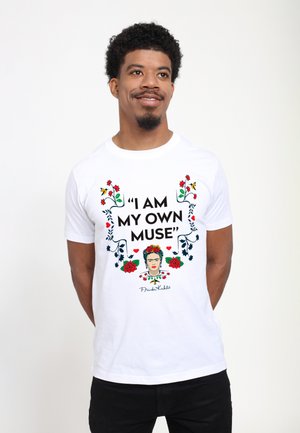 Mand iført hvid t-shirt med teksten "I am my own muse" og Frida Kahlo illustration omgivet af blomster, stående mod en ensfarvet baggrund.