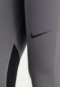 Zbliżenie na szare legginsy sportowe z czarną siateczkową wstawką i czarną logo Nike w kształcie łyżwy na udzie.