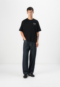 Camiseta de algodón negra con mangas cortas y cuello redondo, que presenta detalles en texto blanco. Combinada con unos vaqueros oscuros de corte holgado y zapatillas negras.