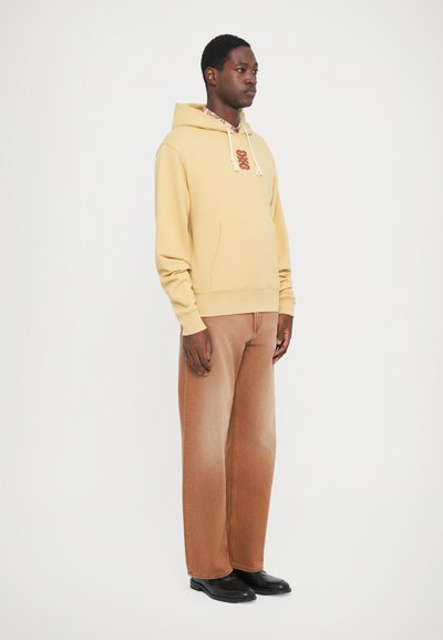 En ung mann i en lys gul hettegenser, brune vide jeans, svarte sko og en mønstret skjorte, står mot en hvit bakgrunn.