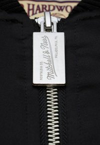 Giacca nera con una zip metallica dotata di un tirante con la scritta "Mitchell & Ness" incisa su un'etichetta rettangolare, circondata da una texture di tessuto morbido.