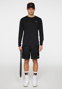 Svart långärmad träningsskjorta med logotyp, svarta shorts, vita sportskor och mönstrade strumpor, håller i ett tennisracket.