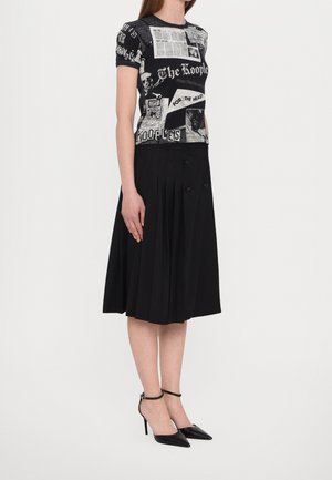 Frau trägt ein schwarzes T-Shirt mit Grafikdruck, einen schwarzen plissierten Midirock mit Knöpfen und schwarze spitze High Heels mit Knöchelriemen.