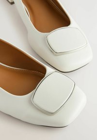 Ballers en cuir blanc avec un bout carré, présentant un grand accent carré en argent à l'avant et une doublure intérieure lisse marron.