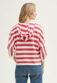 Pullover a righe rosse e bianche con cappuccio, vestibilità rilassata, maniche a tre quarti e tessuto testurizzato. La vista posteriore mostra una cucitura centrale e orlo spaccato.