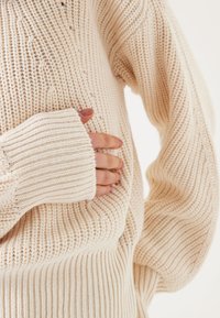 Beige gebreide sweater met geribde manchetten en lijf, met een gestructureerd patroon. Kenmerken zijn een losse pasvorm en een zachte, chunky textuur.