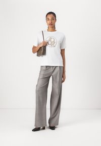 Weißes T-Shirt mit einem runden braunen Grafikdesign, kombiniert mit hellgrauen karierten weiten Hosen und schwarzen High Heels. Hält eine braune Tasche.