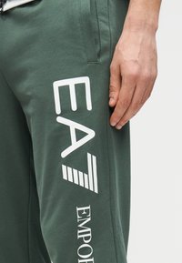 Zelené tepláky z bavlny s bielym grafickým textom "EA" a "EMPORIO" na boku, s jemnou textúrou a voľným strihom.