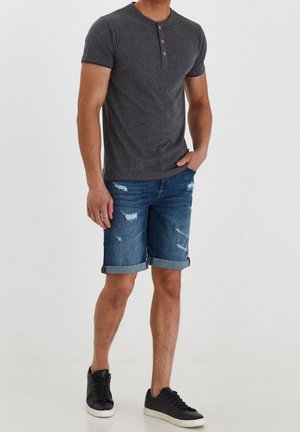 Homme portant un t-shirt henley à manches courtes gris foncé, un short en jean usé et roulé, et des baskets basses noires sur un fond uni.