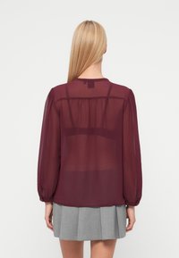 Bordo-teemaline läbipaistev pluus mullitatud pikkade varrukate, volditud seljaga detaili ja ümmarguse kaelusega, mida kannab koos halli volditud seelikuga.