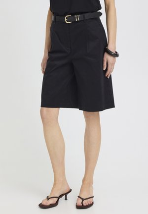 Femeie purtând pantaloni scurți negri până la genunchi, curea neagră, top negru fără mâneci, brățară neagră și sandale negre cu toc, stând în fața unui fundal alb.