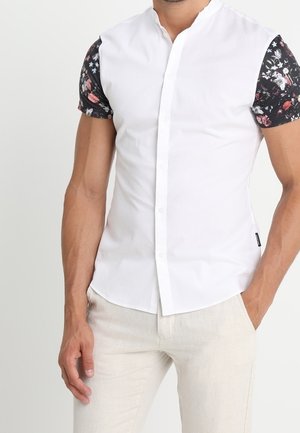 Mężczyzna ubrany w dopasowaną białą koszulę zapinaną na guziki z czarnymi kwiatowymi krótkimi rękawami i jasnobeżowe spodnie, jedną ręką w kieszeni.