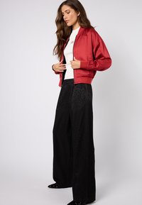 Czerwona satynowa kurtka bomberka z prążkowanymi mankietami, zestawiona z czarnymi spodniami o szerokich nogawkach z delikatnym wzorem w cętki i fakturowaną tkaniną.