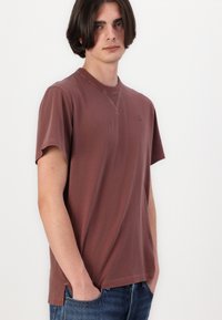 Tricou din bumbac maro cu mâneci scurte, guler rotund și un logo subtil pe piept; asortat cu blugi din denim albastri.