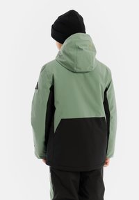 Groene en zwarte, waterdichte jas met een capuchon, voorzien van een grote achterzak en reflecterende merktekens op de bovenrug.